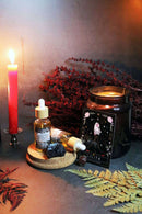Eine Ritual-Altar-Szene zeigt zwei Ritualöl-Flaschen mit Pipette (eine stehend, eine liegend), einen schwarzen Stein, eine "Lilith"-Orakelkarte, ein großes braunes Kerzenglas und eine brennende rote Stabkerze. Rote und grüne Farne, getrocknete Pflanzen und ein Korktablett vervollständigen das magische Arrangement auf einer dunklen Oberfläche.