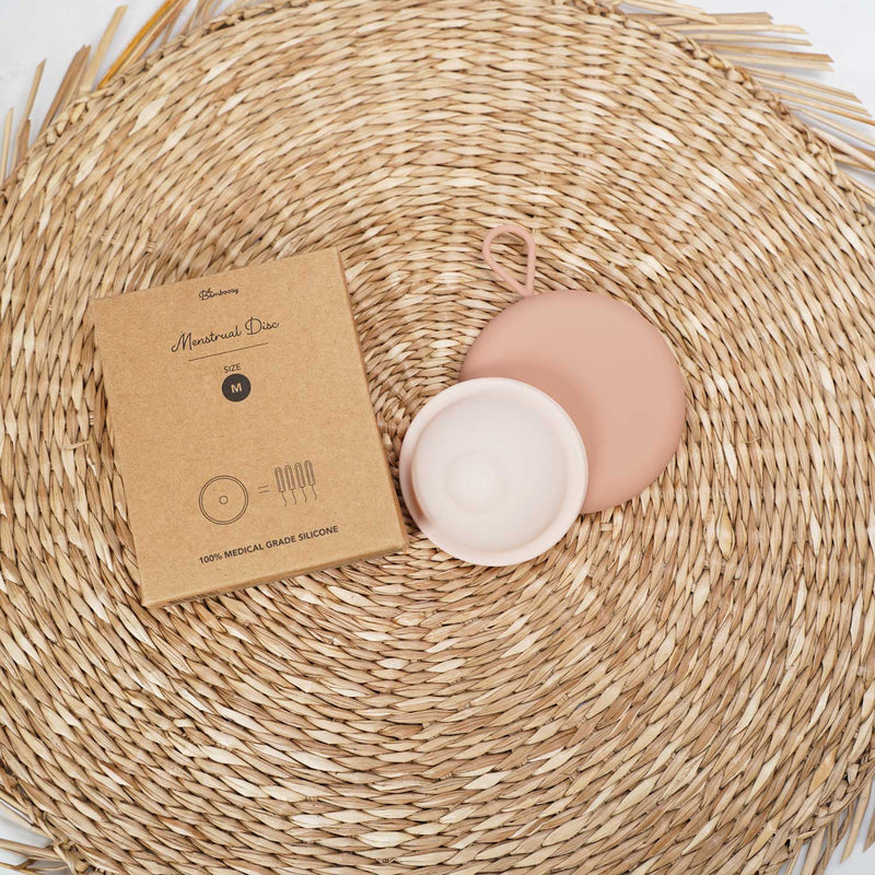 Eine rosa Menstruationsscheibe in Größe M, aus 100% medizinischem Silikon, wird zusammen mit ihrer braunen Kartonverpackung auf einer gewebten Matte präsentiert.