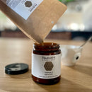 Kräuterpulver wird aus einem Papierbeutel in ein Glasgefäß mit Triphala-Verdauungstonikum gegossen.