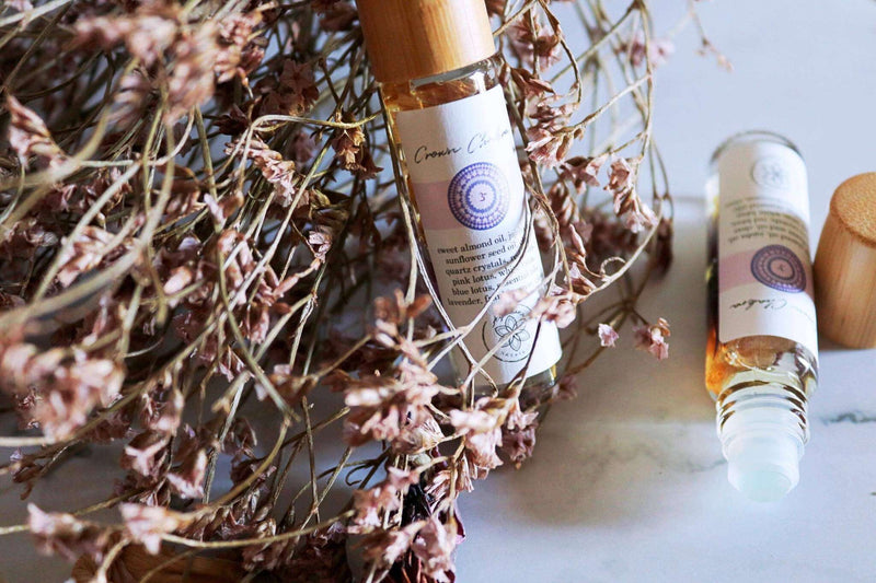 Eine Glas-Roll-On-Flasche mit Bambusdeckel und Aufdruck “Crown Chakra” ruht in einem Geflecht getrockneter rosa Statice-Blüten auf weißem Marmor. Daneben liegt eine zweite Flasche ohne Deckel, die eine Metallkugel und goldfarbenes Öl mit Blüten zeigt. Sanftes Tageslicht wirft zarte Schatten auf Glas und Blüten.