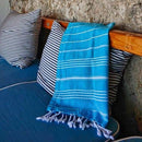 Ein hellblau gestreiftes Handtuch mit weißen Quasten, das über eine Holzbank mit gestreiften Kissen hängt.