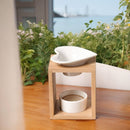 Eine Aromalampe aus Bambusholz mit einer herzförmigen Keramikschale, auf einem Holztisch vor einem Fenster mit Blick auf eine Hafen- oder Wasserlandschaft und umgebendes Grün.