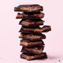 -  Ein hoher Stapel von unregelmäßigen Stücken dunkler Schokolade mit Haselnuss-Füllung, die auf einem rosa Hintergrund übereinander gestapelt sind.