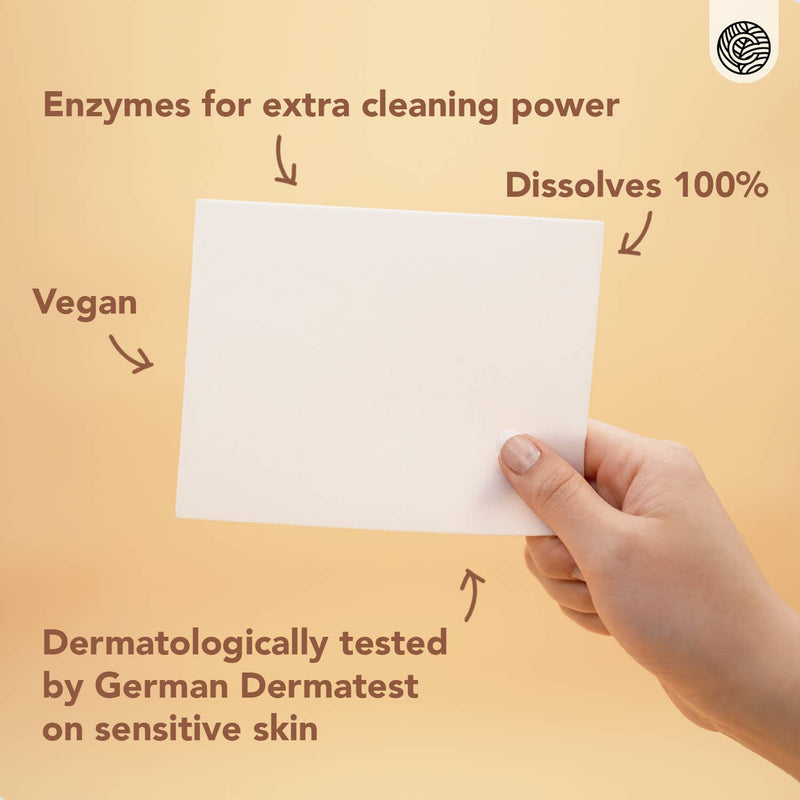 Eine Hand hält ein weißes Waschmittelblatt, mit Text, der seine Eigenschaften hervorhebt: "Enzyme für zusätzliche Reinigungskraft", "Löst sich zu 100% auf", "Vegan" und "Dermatologisch getestet vom deutschen Dermatest an empfindlicher Haut".