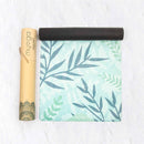 Eine teilweise ausgerollte Myga Yogamatte mit einem hellen botanischen Blattmuster in Blau- und Grüntönen, die neben ihrer hohen, gebrandeten braunen Kartontragrolle liegt.