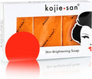 Eine weiße Schachtel Kojie.san Skin Brightening Soap mit einem durchsichtigen Fenster, das drei orangefarbene Seifenstücke im Inneren zeigt, mit dem angeschnittenen Bild eines Frauengesichts und einem auffälligen roten runden Aufkleber in der unteren linken Ecke.