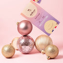 Eine "delikao Feel Good" Schokoladenverpackung lehnt an einem Stapel roségoldener Weihnachtskugeln, während eine weitere Kugel oben balanciert. Der Hintergrund ist rosa, mit weiteren Weihnachtskugeln und einem angedeuteten zweiten Schokoriegel.