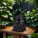 Ein beleuchteter Buddha-Zimmerbrunnen auf einem Holztisch, umgeben von üppig grünen Pflanzen.