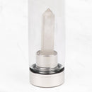 Eine Nahaufnahme des unteren Teils einer durchsichtigen Glaswasserflasche, die den Edelstahlsockel und einen prominenten, klaren Quarzkristall zeigt, der von diesem nach oben ragt. Die Flasche steht auf einer weißen Marmoroberfläche.