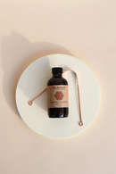 Eine braune PAAVANI Zimt-Ölziehe-Flasche ist mittig auf einer runden weißen Plattform platziert, mit einem kupferfarbenen Zungenreiniger, der darum geschlungen ist. Die Objekte werfen weiche Schatten auf einem hellen Pfirsich-Hintergrund.