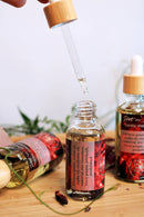 Eine Hand hält einen Tropfer über einer offenen Flasche "Hot n Spicy Tantric Massage Oil", bereit, die klare, mit Pflanzen angereicherte Flüssigkeit abzugeben. Zwei weitere Flaschen desselben Öls sind auf der Holzoberfläche sichtbar, begleitet von verstreuten getrockneten Blütenblättern und grünem Laub, vor einem weichen weißen Hintergrund.