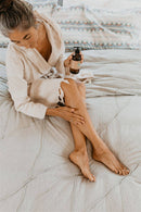 Eine Frau mit ergrautem Haar, bekleidet mit einem weiß gestreiften Bademantel, sitzt auf einem Bett und trägt mit ihrer rechten Hand Körperöl auf ihr Bein auf, während sie die PAAVANI Vata Körperölflasche in ihrer linken Hand hält.