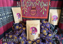 Eine Präsentation von drei Ancient Purity Blaue Lotusblume Produktbeuteln inmitten einer großen Menge loser, getrockneter lila-goldener Blüten. Der Hintergrund zeigt ein farbenprächtiges, traditionelles Textil mit geometrischen Mustern.