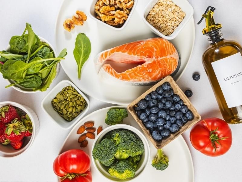 Gesunde Lebensmittel wie Lachs, Beeren, Brokkoli, Nüsse und Olivenöl auf weißem Hintergrund.