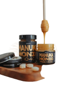 Goldener honig tropft von einem holzlöffel über zwei schwarz etikettierten anuk manuka honey mgo400+ gläsern mit weißen tabletten daneben
