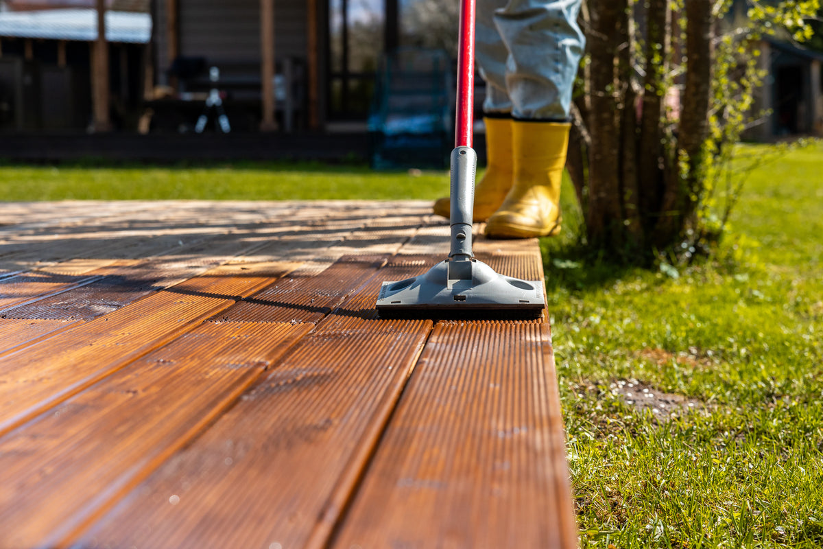 Person reinigt eine Holzterrasse im Garten mit einem Bodenwischer und gelben Gummistiefeln.