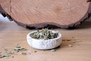 Eine weiße Steinschale, gefüllt mit getrockneten grünen und braunen Blättern, steht auf einem hellen Holztisch. Ein großes Stück eines Baumstamms ist im Hintergrund zu sehen. Einige der Blätter sind um die Schale herum auf dem Tisch verstreut.