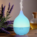 Ein elektrischer, blau-hellblauer tropfenförmiger Aroma-Diffusor, der Nebel ausstößt und blau leuchtet, neben Lavendelblüten und einer Flasche ätherischen Öls.