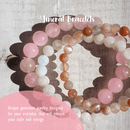 Eine Sammlung von Edelsteinarmbändern in Rosa, Weiß und Braun auf einer weißen Holzoberfläche mit Text.