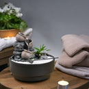 Ein Desktop-Zen-Garten mit einem stufigen Wasserbrunnen und weißem Sand auf einem Tisch mit Kerzen und Handtüchern.