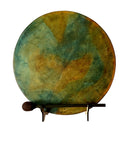 Eine runde Handtrommel (Rahmen- oder Schamanentrommel) mit einem bemalten Fell in auffälligen, gesprenkelten Petrol-/Grün-, Gold- und Orangetönen, die auf einem kleinen Holzständer ruht, mit einem dunklen Holz-Schlägel davor platziert.