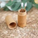 Zwei wiederverwendbare Wattestäbchen mit einer strukturierten und einer spitzen Silikonspitze, jeweils in einem geöffneten Bambusbehälter auf einer geflochtenen Matte mit verschwommenem grünem Hintergrund.