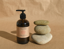 Eine braune Glasflasche mit Pumpspender für Kapha-Körperöl neben einem Stapel aus drei glatten Flusssteinen auf braunem Papierhintergrund.