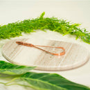 Ein Zungenreiniger aus Kupfer liegt auf einer ovalen Marmorplatte, umgeben von grünen Blättern.