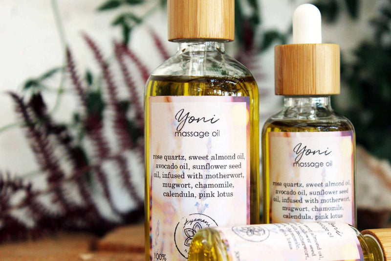 Drei Glasflaschen mit Pipette für "Yoni Massage Oil" mit Bambusverschlüssen sind auf einem Baumstumpf ausgestellt. Eine Flasche liegt auf der Seite, während zwei aufrecht stehen und unterschiedlich groß sind. Das klare Öl enthält infundierte Pflanzenstoffe, und unscharfes grünes und rotes Laub ist im Hintergrund zu sehen.