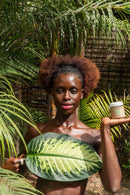 Eine Frau mit braunen Locken steht in einem tropischen Garten, hält ein großes grünes Blatt vor ihrem Körper und präsentiert in der anderen Hand einen Tiegel PAAVANI Vata Cleanser & Mask.