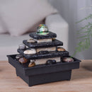 Ein mehrstufiger Zen-Steinbrunnen mit einer leuchtenden Glaskugel und dekorativen Flusssteinen, platziert auf einer Holzoberfläche in einem Wohnzimmer.