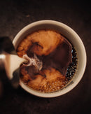 Cremige Milch wirbelt in dunklen Kaffee, wodurch eine künstlerische Mischung aus hellbraunen und tiefschwarzen Farbtönen in einer weißen Keramiktasse entsteht, mit zarten Blasen, die sich auf der Oberfläche bilden. Diese Draufsicht fängt den Moment des Mischens ein und betont die reichen Texturen und einladende Wärme.