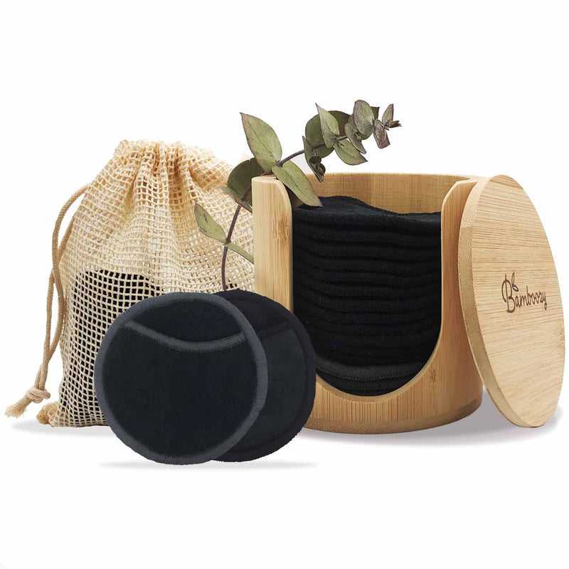 Set wiederverwendbarer schwarzer Make-up-Entfernerpads in einem Bambushalter und einem Wäschenetz, begleitet von einem kleinen grünen Pflanzenzweig.
