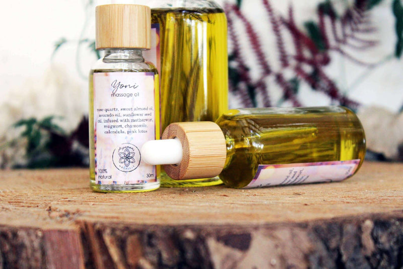 Drei Glasflaschen mit Pipette für "Yoni Massage Oil" mit Bambusverschlüssen sind auf einem Baumstumpf ausgestellt. Eine Flasche liegt auf der Seite, während zwei aufrecht stehen und unterschiedlich groß sind. Das klare Öl enthält infundierte Pflanzenstoffe, und unscharfes grünes und rotes Laub ist im Hintergrund zu sehen.