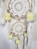 Eine detaillierte Nahaufnahme eines dreifachen Traumfängers mit weißen Häkelspitzenmustern in beige-farbenen Reifen, verziert mit gelben Pompons und weißen Quasten.