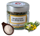 Ein Glasgefäß mit der Aufschrift „Bergkernsalz mit Wildkräutern“ und Kräutern darin, verschlossen mit einem Metalldeckel. Daneben stehen ein Holzlöffel mit Salz und eine Schale mit bunten Kräutern.