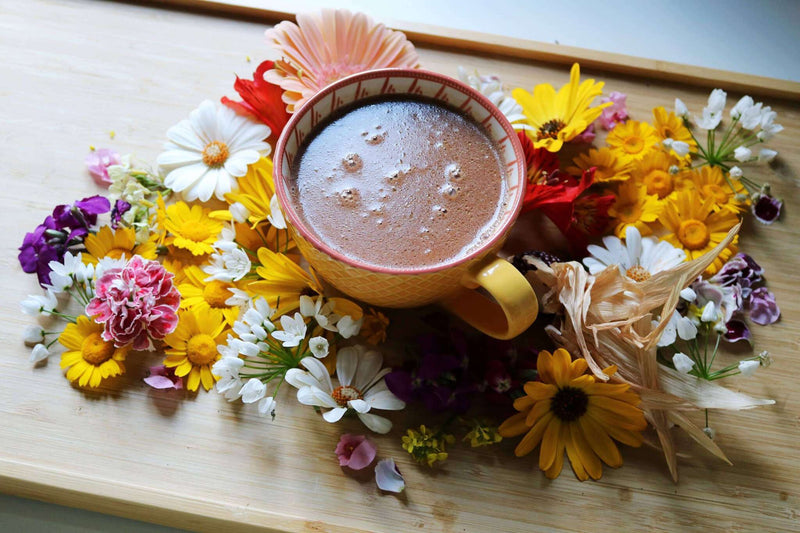Eine farbenfrohe Auswahl frischer Blumen in Rosa-, Gelb-, Weiß- und Lilatönen umgibt eine orangefarbene Keramiktasse mit einem schaumigen Getränk. Alle Elemente sind kunstvoll auf einem hellen Holztablett arrangiert, was eine lebendige und ansprechende Darstellung schafft.