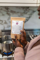 Eine Person hält eine Packung „PAAVANI HERBAL YONI STEAM“ über einem Topf mit kochendem Wasser auf einem Herd.