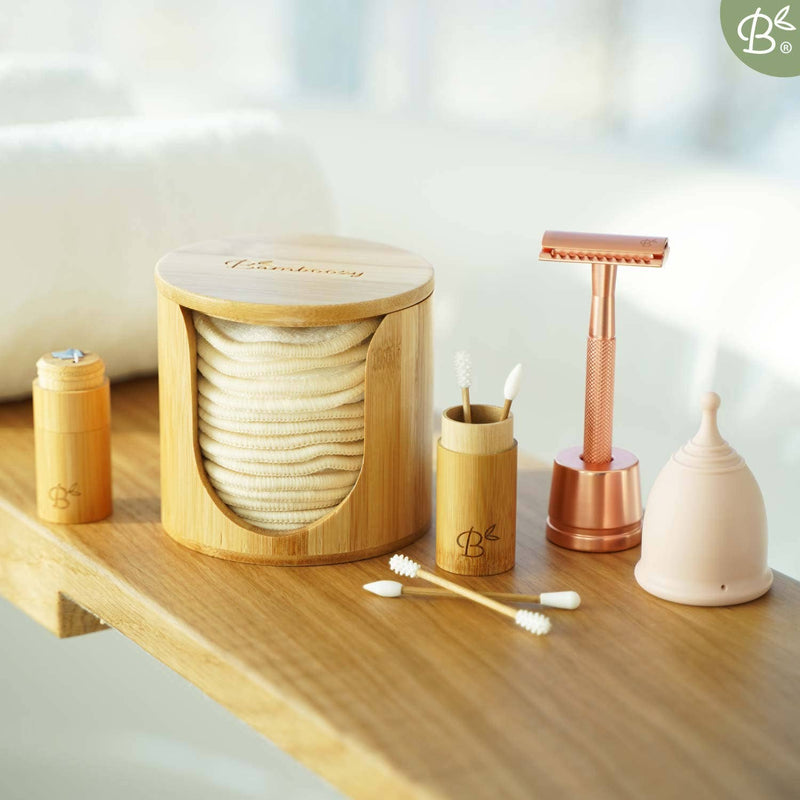 Eine umfassende Darstellung von Bamboozy Körperpflegeprodukten: ein Bambushalter mit wiederverwendbaren Wattepads, Bambusbehälter für Wattestäbchen und Zahnseide, ein roségoldener Rasierhobel und eine rosa Menstruationstasse, alles auf einem Holzregal.