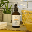  Eine dunkle Glasflasche "Comptoir des Huiles Huile de Massage Visage" mit einem Pumpspender, für reife Haut, steht auf einer Arbeitsplatte mit weißen Fliesen im Hintergrund und einer grünen Pflanze links.