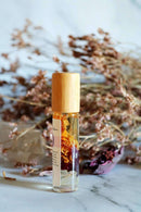  Eine Roll-on-Flasche mit Chakra-Öl und einem Holzdeckel steht auf einem leuchtend blauen und lila Fluoritkristall. Im Hintergrund sind getrockneter Lavendel und braune Pflanzen zu sehen.