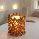 Nahaufnahme eines rustikalen Kerzenhalters aus ineinandergreifenden Holzstäbchen, der auf einem weißen Tisch warm leuchtet.