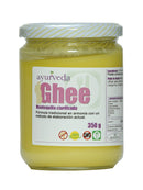 Ein Glasgefäß mit "Ayurveda autentico Ghee" (geklärte Butter) mit gelb-weiß gestreiftem Deckel. Das violette Vorderetikett trägt die Aufschrift "Mantequilla clarificada", mit Symbolen für "BIO", "100% Natural", "Sin Gluten", "OMG" (gentechnikfrei) und "350 g".