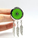 Ein silberfarbener Aromadiffusor-Anhänger im Traumfänger-Stil mit einem floralen Ausschnitt über einem leuchtend neongrünen Filzpad und drei herabhängenden silbernen Feder-Anhängern, der zwischen zwei Fingern gehalten wird.