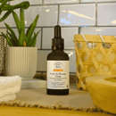 Eine dunkle Glasflasche "Comptoir des Huiles Huile de Massage Visage" mit Pipette, für reife Haut, steht auf einer Arbeitsplatte mit weißen Fliesen im Hintergrund und einer grünen Pflanze links.