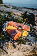Gerollte orange und gelb gestreifte Handtücher mit Quasten auf einem steinigen Strand.