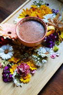 Eine lebendige Ansammlung frischer Blumen in verschiedenen Farben wie Pink, Gelb, Weiß und Lila ist kunstvoll um eine orangefarbene Keramiktasse mit einem schaumigen Getränk arrangiert. Alle Gegenstände stehen auf einem hellen Holztablett, was eine ruhige und natürliche Ästhetik suggeriert.