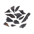 Eine Aufnahme von oben, die eine verstreute Gruppe von etwa einem Dutzend roher, rauer schwarzer Kyanit-Kristallblätter zeigt. Die faserigen, fächerförmigen Kristalle liegen auf einem weißen Hintergrund.