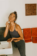 Eine Frau mit aufgetragener Gesichtsmaske, die Augen zufrieden geschlossen, hält eine kleine braune Schale in ihrer linken Hand und trägt die Maske sanft mit ihrer rechten Hand auf ihr Gesicht auf. Orangefarbene Handtücher und ein dekorativer Wandschmuck sind im Hintergrund zu sehen.