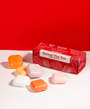 Sechs bunte Seifen liegen auf einer weißen Fläche neben einer roten Box mit der Aufschrift „Reveal The You“. Der kräftige rote Hintergrund vermittelt Lebendigkeit.